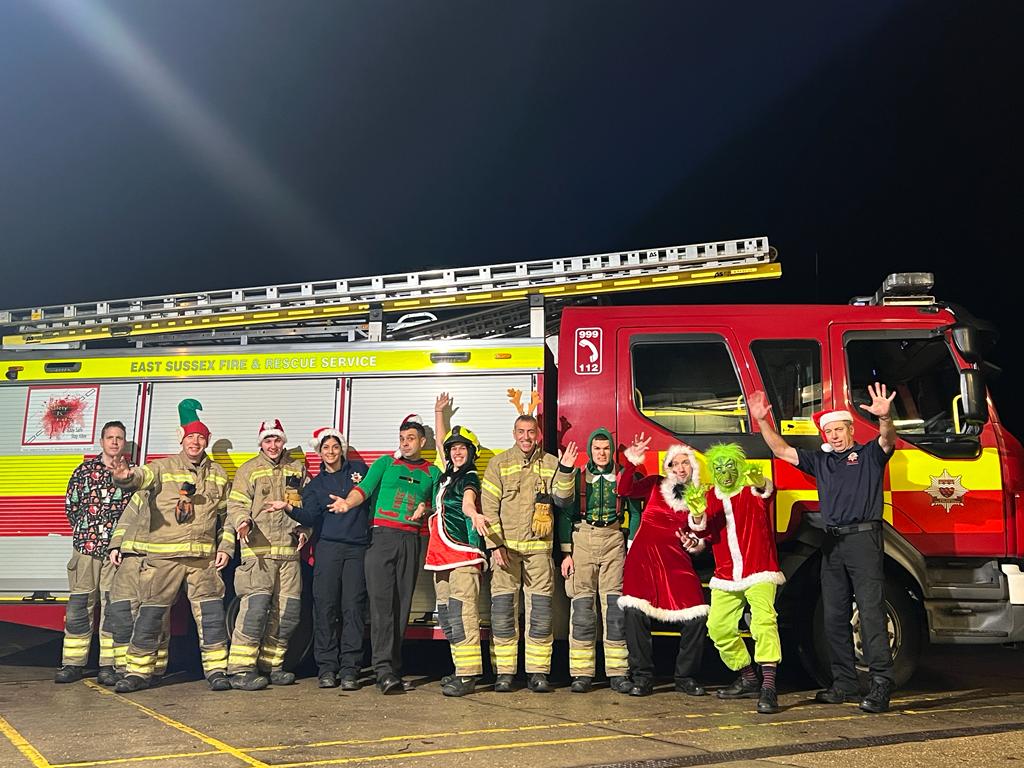 Photo of firefighters in front of fire engine some dressed in festive clothing and someone dressed as a Grinch
