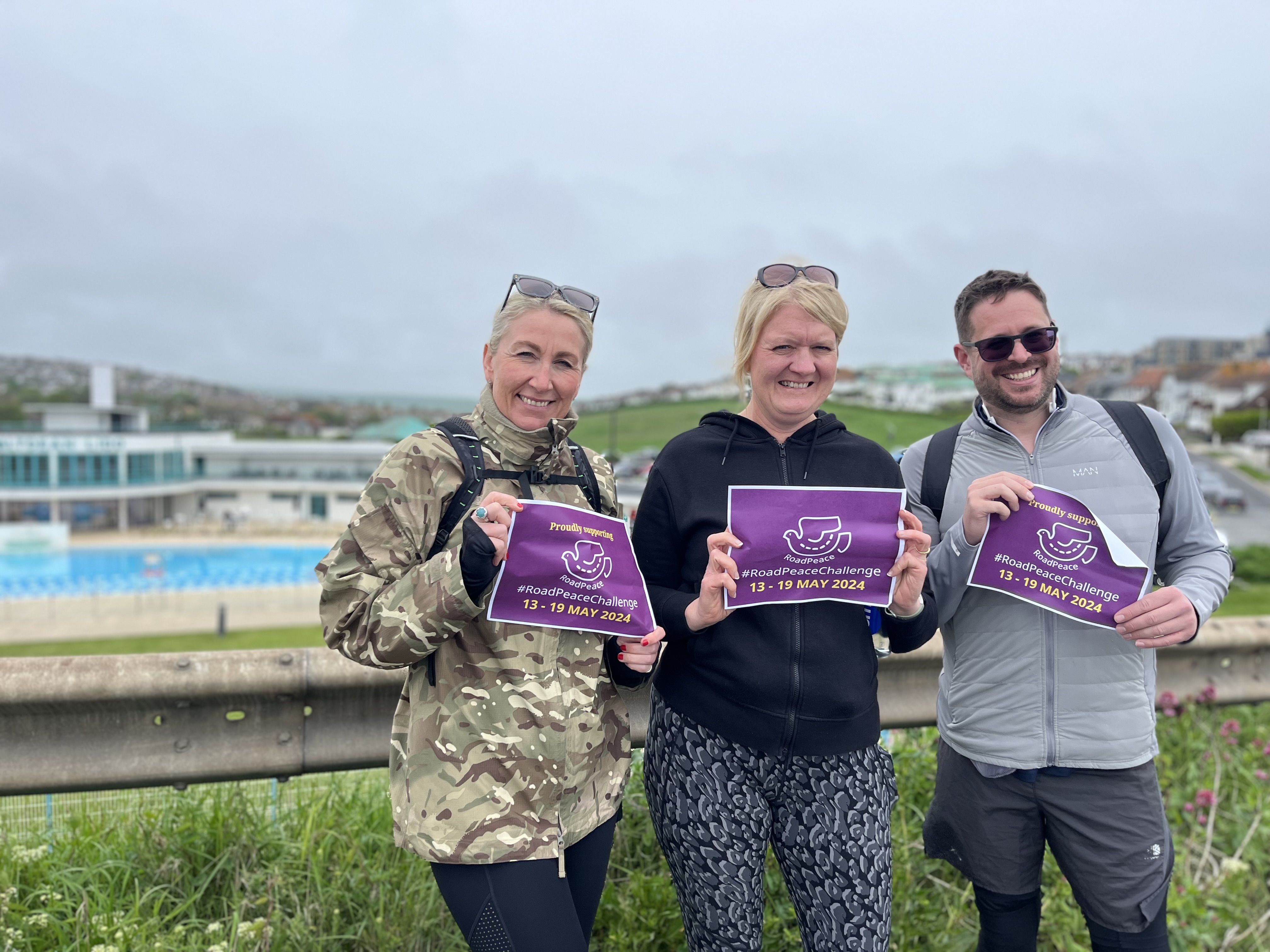 3 people holding a RoadPeace Challenge sign with Saltdean Lido in the background