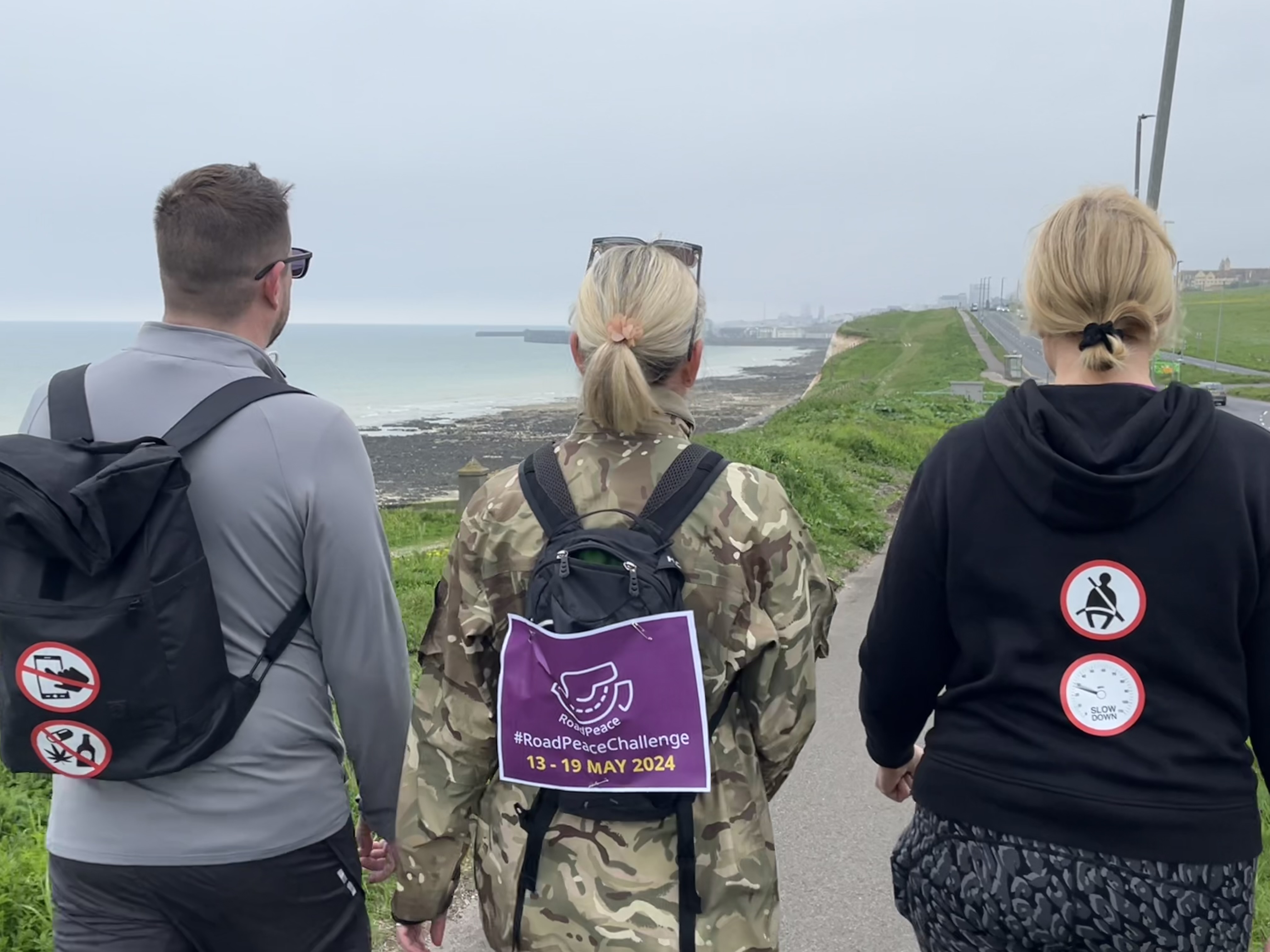 Three people walking with Brighton seafront in the distance. Wearing road safety stickers and a RoadPeace Challenge sign