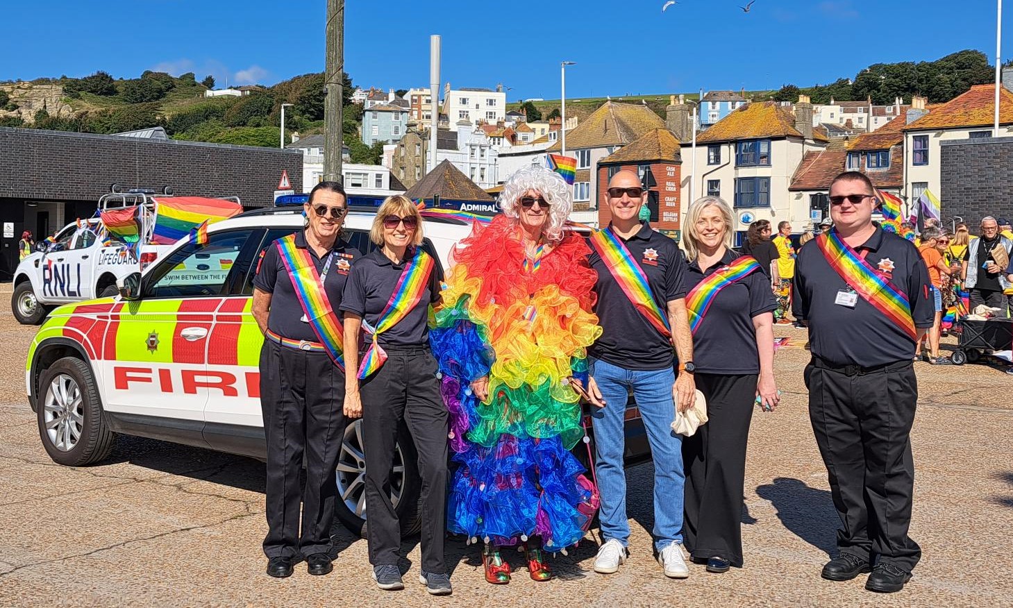 Photo of 6 people in front of fire car wearing pride sashes. Person in the middle is in rainbow coloured feathers. For Hastings Pride 2023