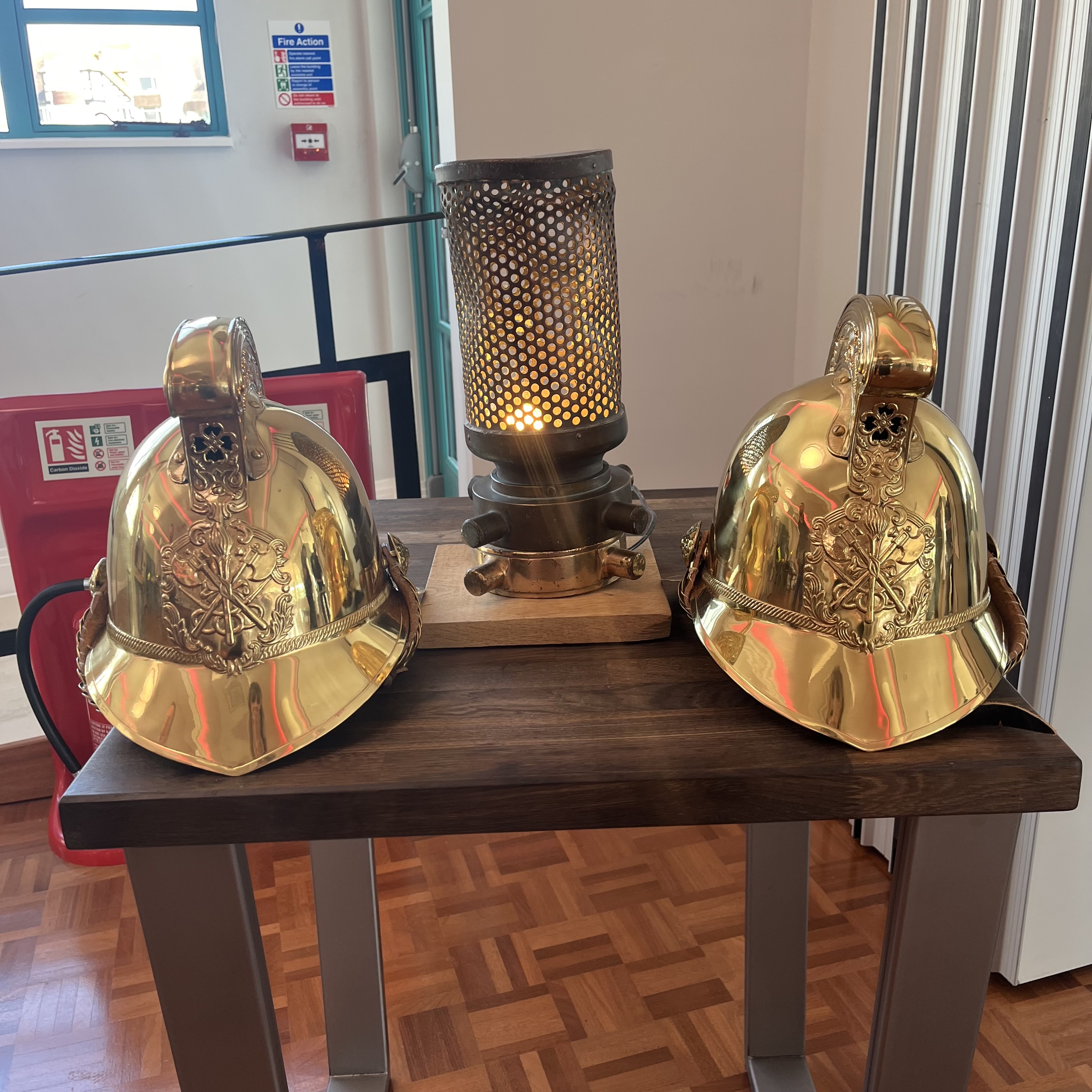 Two old fashioned bronze gold fire helmets on disaplay on wooden table with old fashioned light in between them
