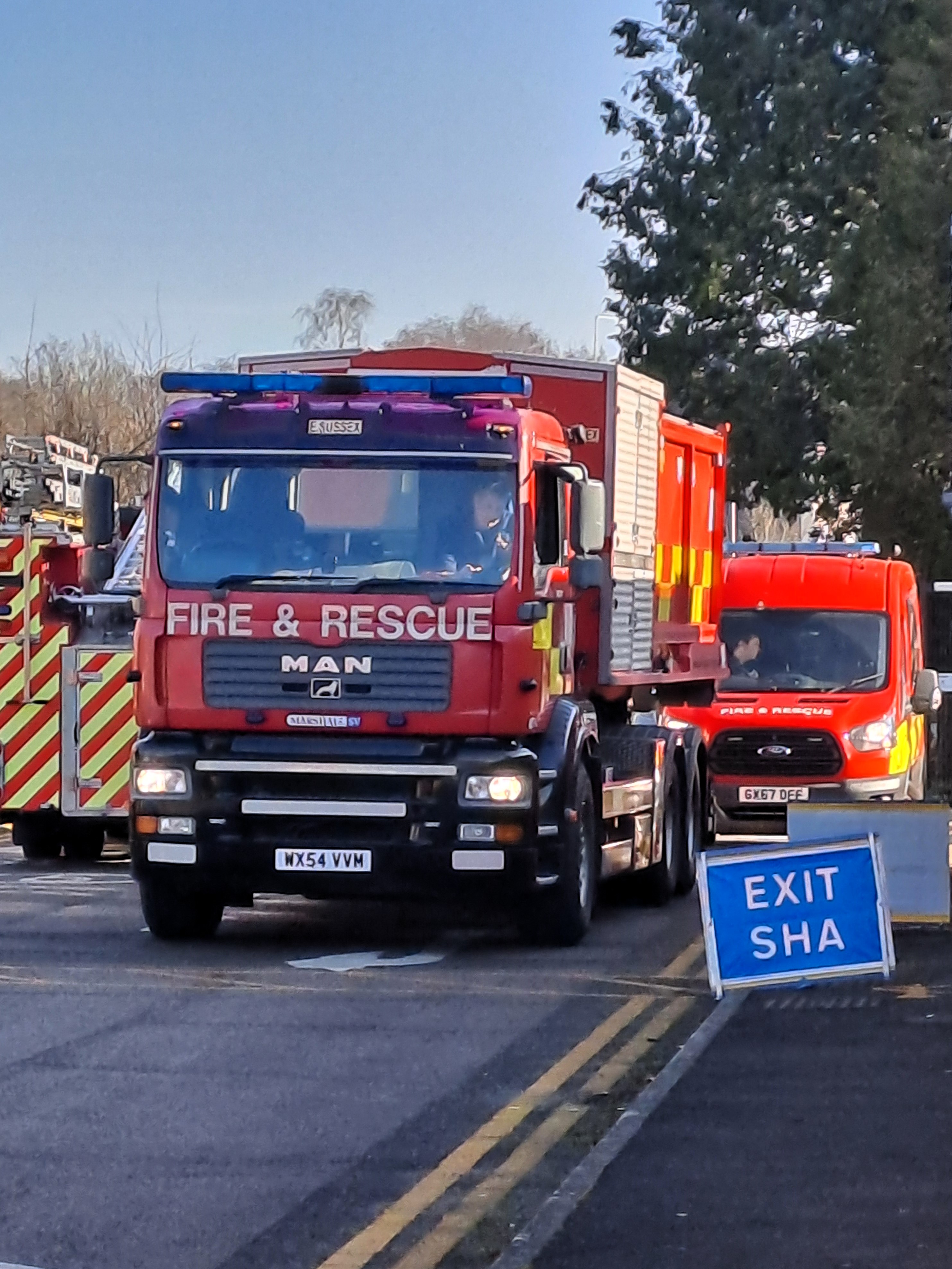 Photo of red fire engine and red fire van on road with blue sign on road that reads EXIT SHA (Strategic Holding Area)