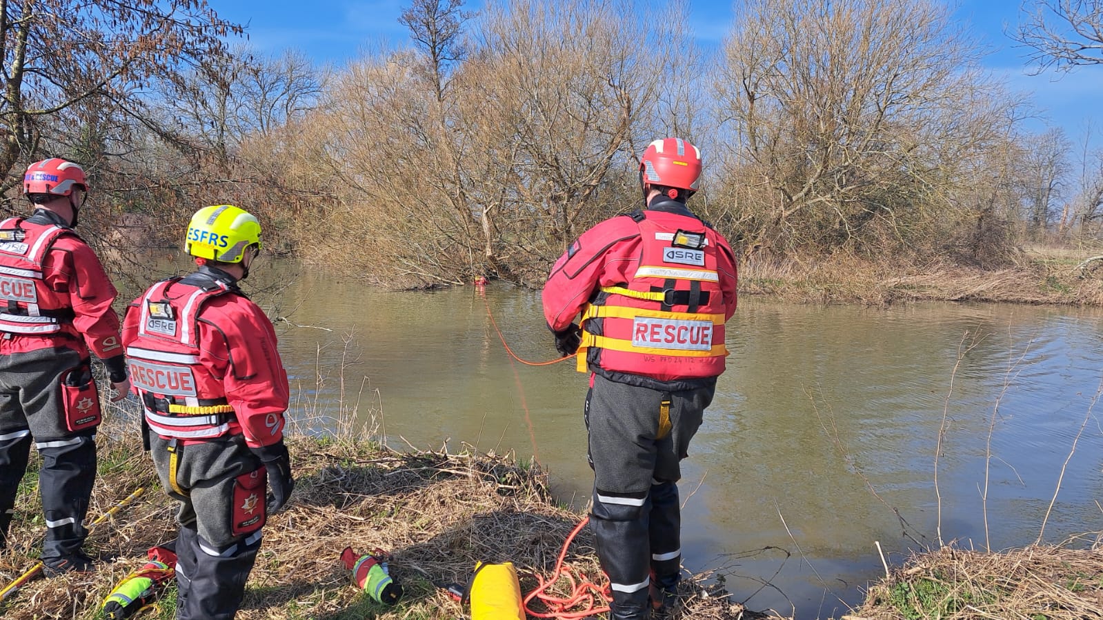 A water rescue crew of three people. Two people in red helmets and one in bright hi viz yellow helmet looking out to a lake or river.