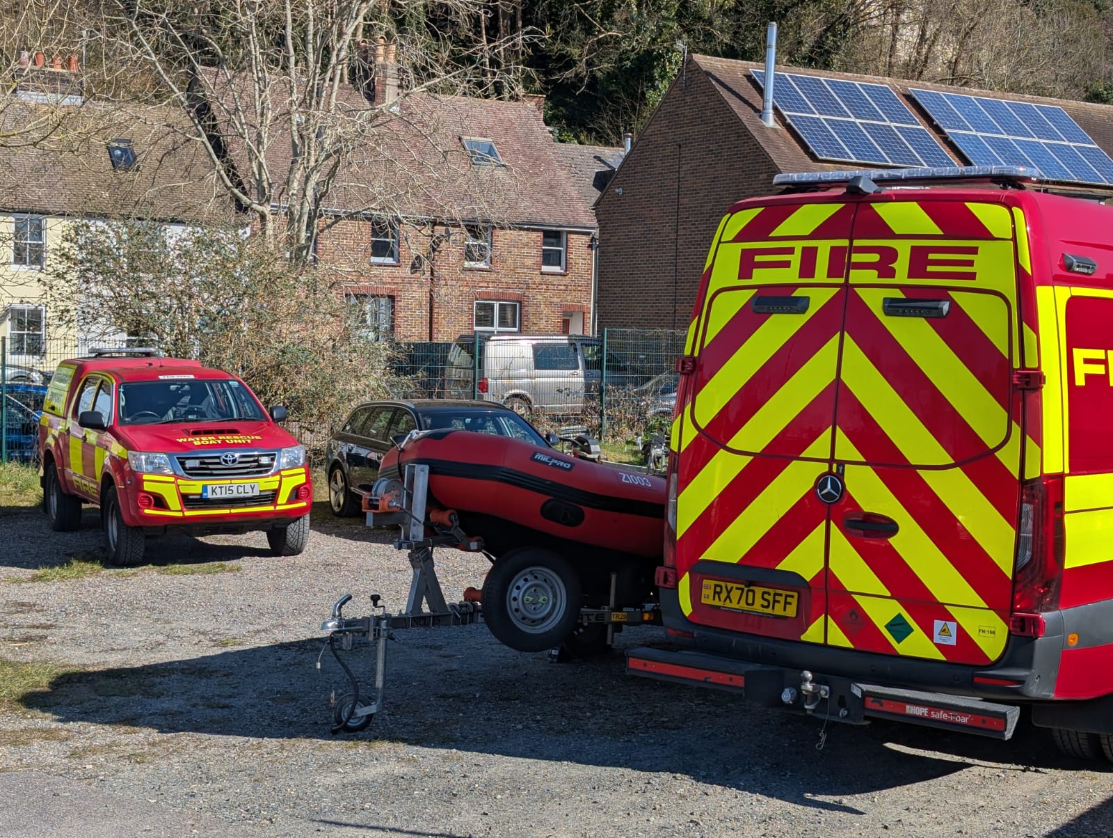 Fire and rescue van with red and yellow chevrons on the back. A red water rescue boat and red landrover in front of some housing.