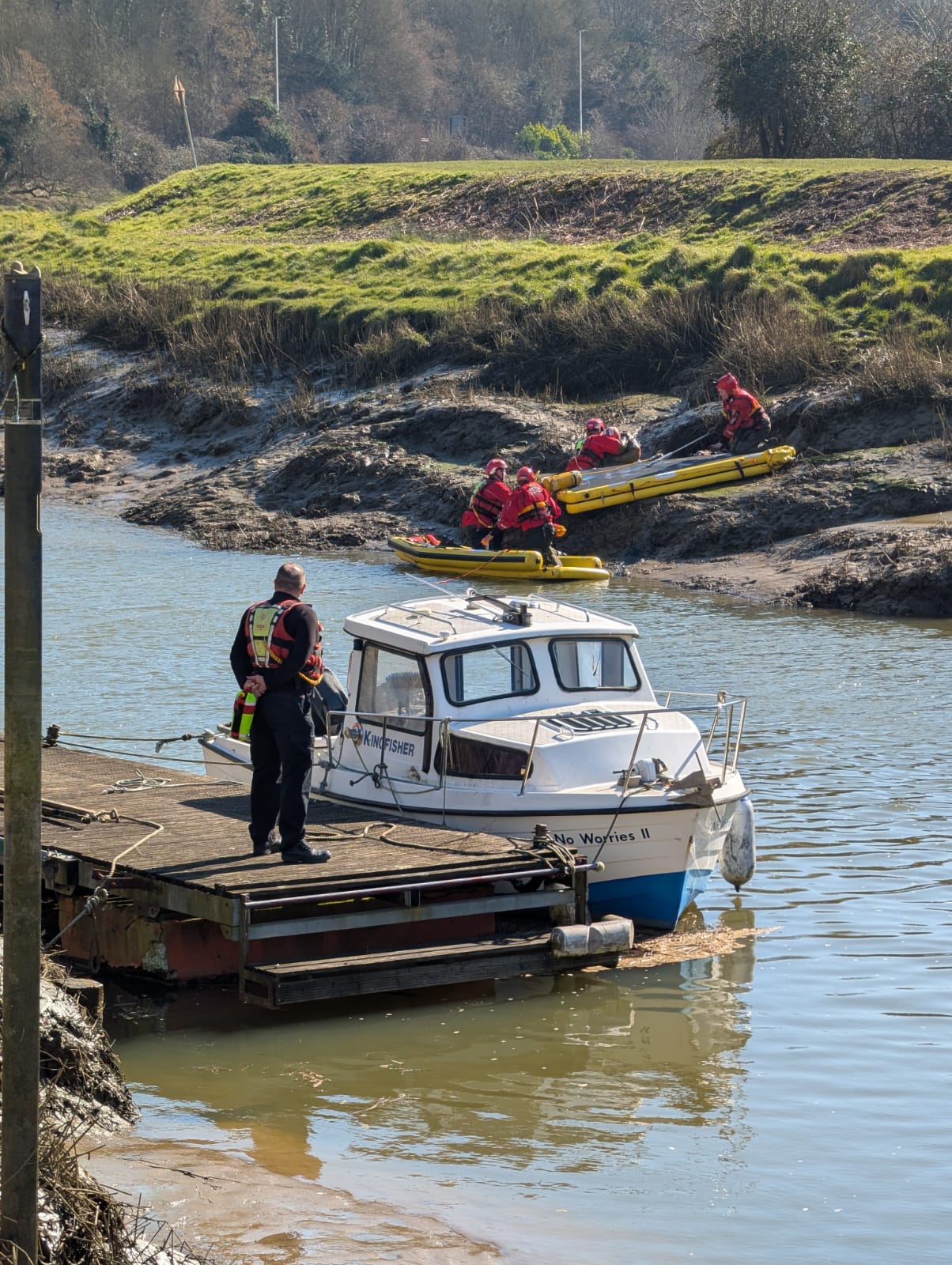 Two people standing by river in front of a small white boat. In the backgrond on a muddy bank are a water rescue crew in red uniform with yellow kayaks.