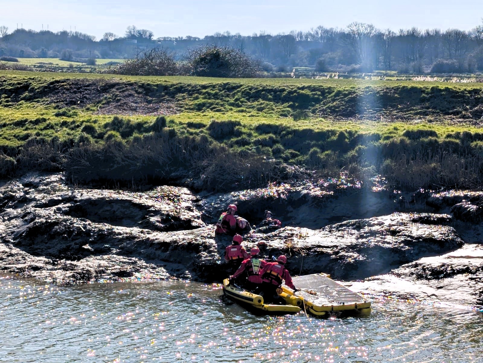 A muddy river bank on a sunny day with fire and rescue crews performing a rescue scenario.