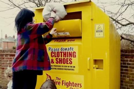 Person putting a bag into The Fire Fighters Charity yellow clothing bank. Two children beside the person. 
