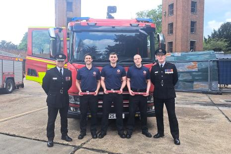 Photo of Fire Service training ground with towers in the background. In front of a red fire engine are 5 people. One uniformed officer - Assistant Director -  on the left black suit, white shirt, black tie and cap with medals on jacket, three firefighters in the middle in navy blue polo shirts and trousers, and one unformed officer - Chief Fire Officer - on the right in black suit, white shirt, black tie and cap with medals on jacket.