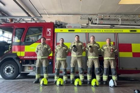Five firefighters standing in front of a fire engine. Their helmets are on the floor in front of each firefighter - four yellow and one white helmet.
