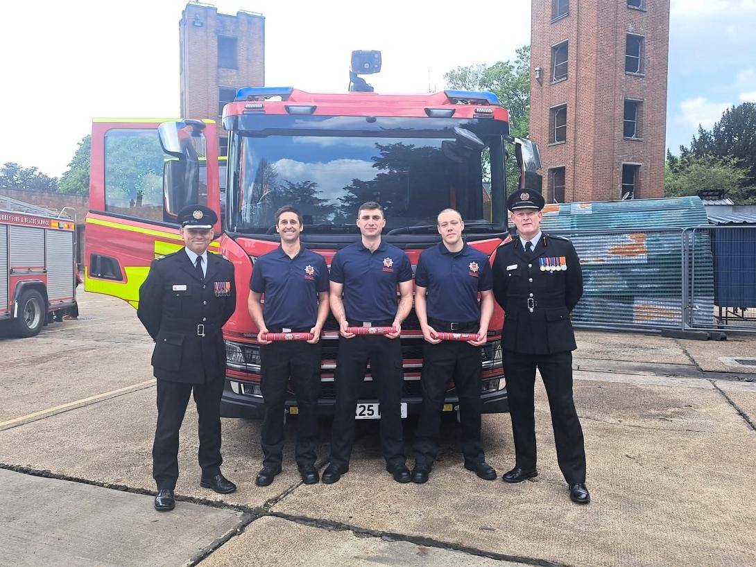 Photo of Fire Service training ground with towers in the background. In front of a red fire engine are 5 people. One uniformed officer - Assistant Director - on the left black suit, white shirt, black tie and cap with medals on jacket, three firefighters in the middle in navy blue polo shirts and trousers, and one unformed officer - Chief Fire Officer - on the right in black suit, white shirt, black tie and cap with medals on jacket.