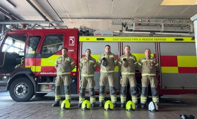 Five firefighters standing in front of a fire engine. Their helmets are on the floor in front of each firefighter - four yellow and one white helmet.